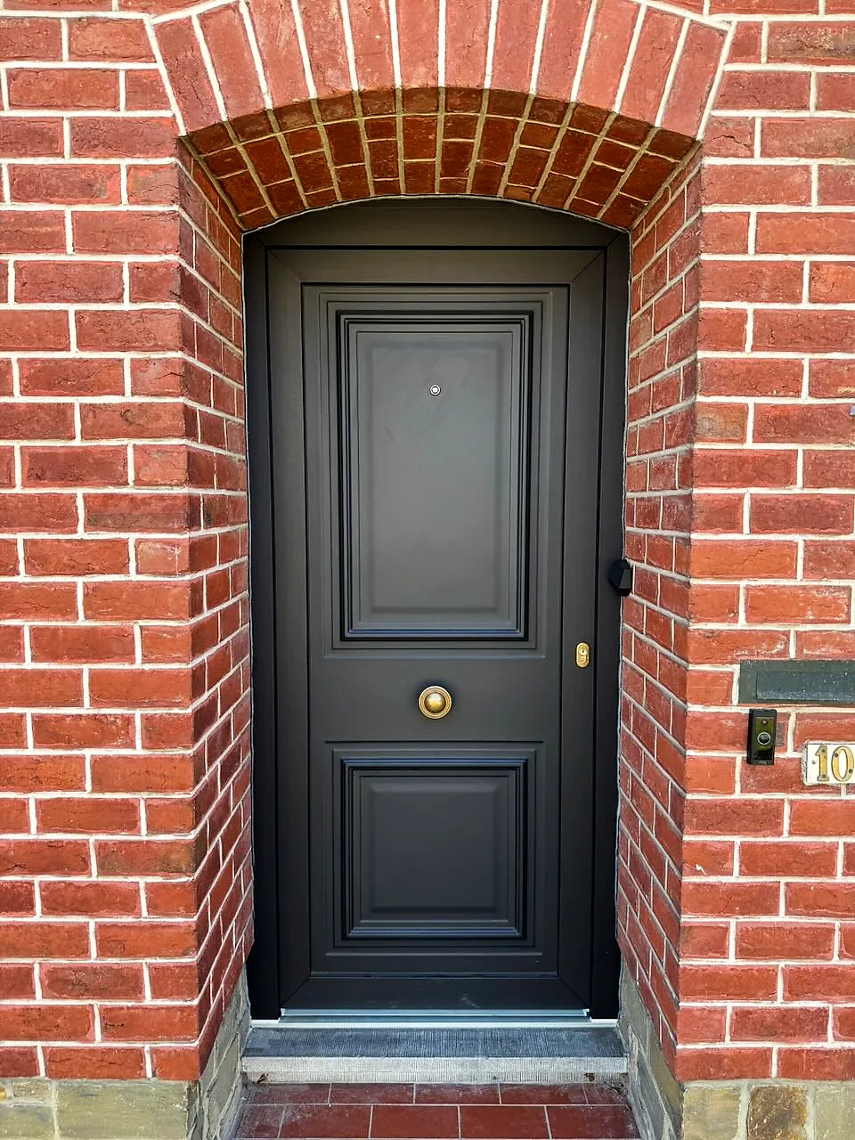 Porte d’entrée anthracite à panneaux moulurés et bouton en laiton, encadrée d’une arche en briques rouges — réalisation Pulsar Windows