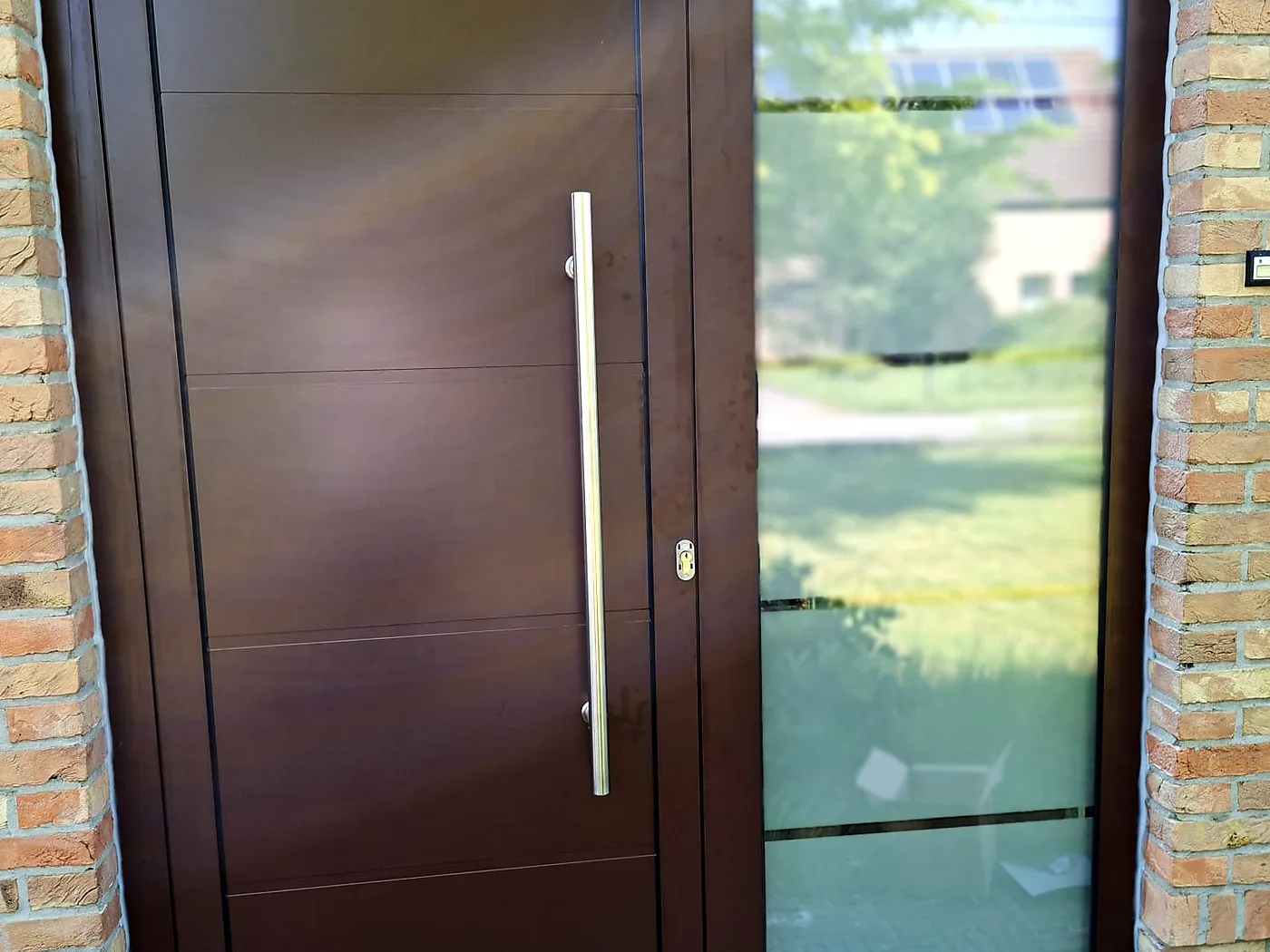 Brown entrance door with modern lines, a long stainless-steel handle and a side glazing panel opening onto the garden — a Pulsar Windows project