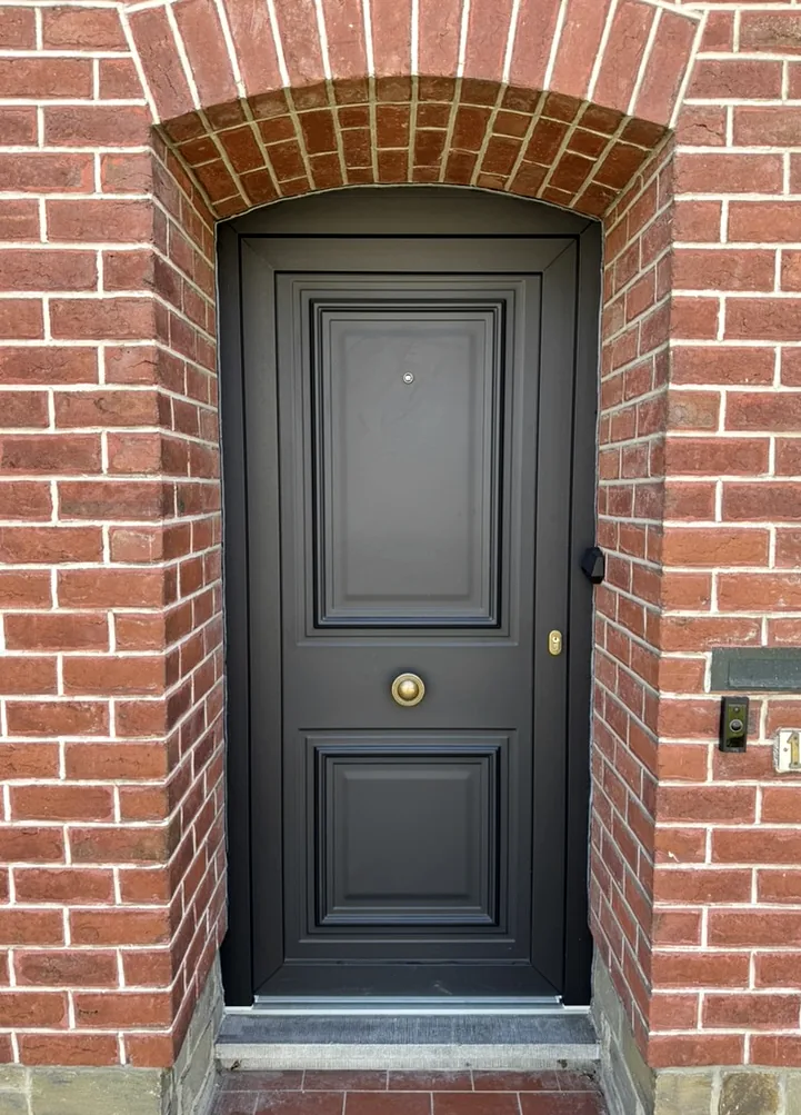 Porte d’entrée noire à panneaux moulurés installée sous un arc en briques.