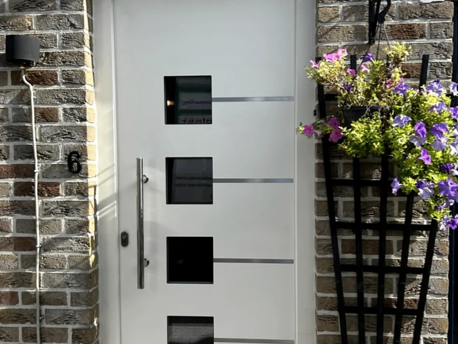 Porte d’entrée gris clair avec quatre inserts vitrés carrés sur une façade en briques.