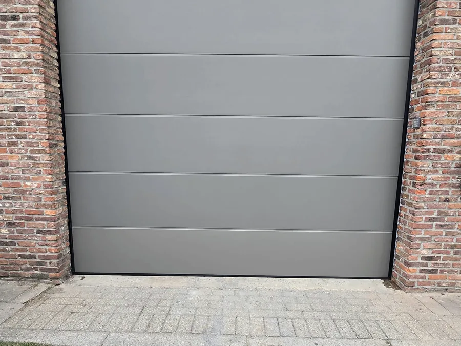 Porte de garage sectionnelle gris clair installée dans un encadrement en briques rouges.